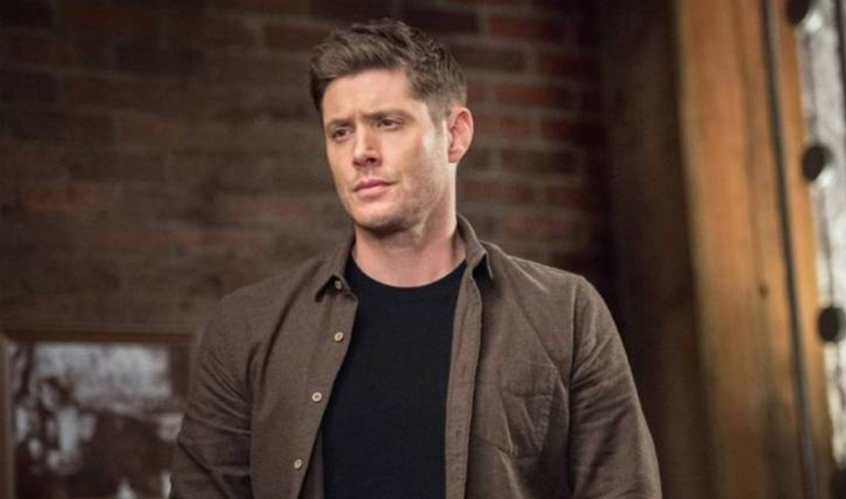 O personagem Dean com uma blusa preta e uma jaqueta marrom na frente d euma parede de tijolos.