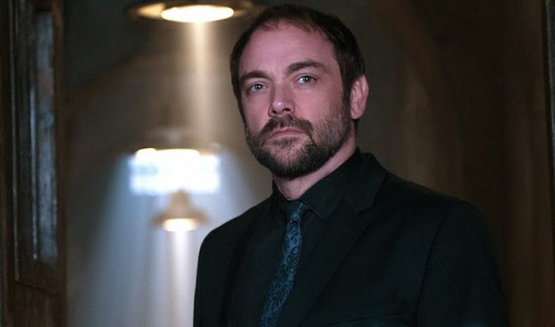 O personagem Crowley vestindo um terno preto e gravata azul.