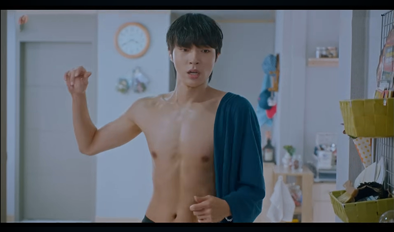 Jovem sem camisa, surpreso, segura uma blusa azul no ombro em um quarto iluminado e organizado com itens pessoais ao fundo.