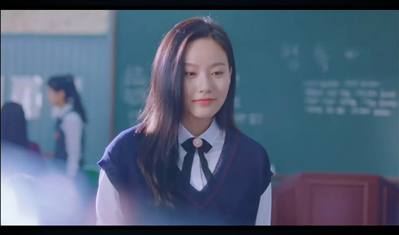 Jovem de uniforme escolar sorri levemente em sala de aula. Usa colete azul-marinho com laço preto e está diante de um quadro verde.