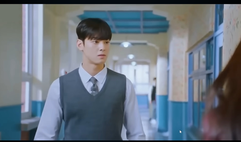 Jovem de uniforme escolar caminha por um corredor, com expressão séria. Ele usa colete cinza sobre camisa branca e gravata. Cena clara e escolar.