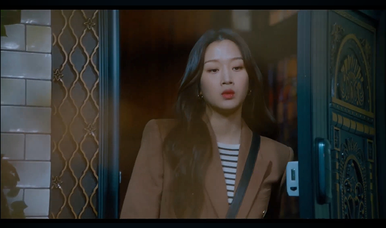 Mulher com expressão séria aparece à porta, usando blazer marrom e blusa listrada. A cena tem iluminação suave e clima dramático.